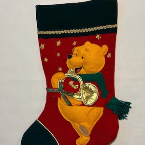 May include: Red Christmas stocking featuring Winnie the Pooh holding a golden bell. The stocking has a green cuff and toe, with gold trim and star accents. A green scarf and bow adorn the bell.