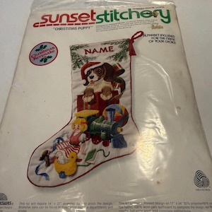 May include: A Sunset Stitchery Christmas Puppy stocking kit. The design features a puppy wearing a Santa hat, surrounded by Christmas toys. The kit includes an alphabet for personalization and measures 17" x 24" when completed.
