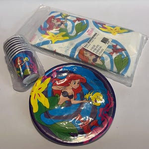 May include: A set of The Little Mermaid party supplies, including a tablecloth, paper plates, and cups. The tablecloth is white with a blue and green pattern featuring Ariel, Flounder, and Sebastian. The paper plates are purple with a blue and green pattern featuring Ariel and Flounder. The cups are purple with a blue and green pattern featuring Ariel and Sebastian.