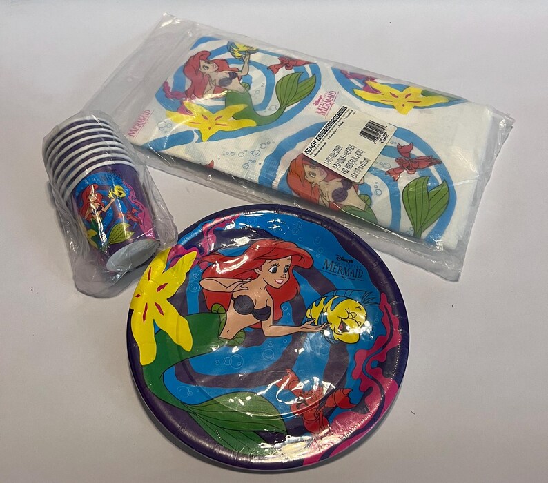 May include: A set of The Little Mermaid party supplies, including a tablecloth, paper plates, and cups. The tablecloth is white with a blue and green pattern featuring Ariel, Flounder, and Sebastian. The paper plates are purple with a blue and green pattern featuring Ariel and Flounder. The cups are purple with a blue and green pattern featuring Ariel and Sebastian.