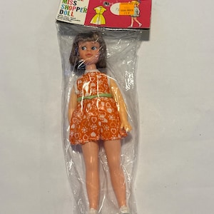 May include: A vintage Little Miss Shopper Doll in its original packaging. The doll has brown hair and is wearing an orange dress with white heart-shaped patterns and a green trim. The packaging includes the doll's name and a picture of a dress.
