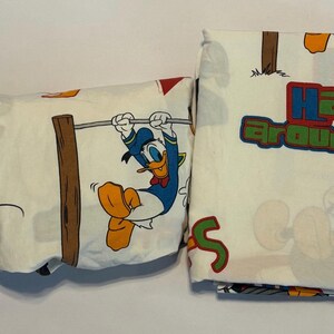 Puede incluir: S&aacute;banas blancas con los personajes de dibujos animados Donald Duck y Mickey Mouse. Donald Duck se balancea en una cuerda y Mickey Mouse est&aacute; de pie. Las palabras "Hang around" est&aacute;n impresas en colores.