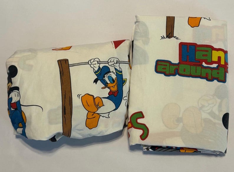 Puede incluir: S&aacute;banas blancas con los personajes de dibujos animados Donald Duck y Mickey Mouse. Donald Duck se balancea en una cuerda y Mickey Mouse est&aacute; de pie. Las palabras "Hang around" est&aacute;n impresas en colores.