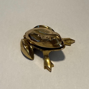 Pode incluir: Uma estatueta de rã de latão com uma superfície detalhada e texturizada. A rã está sentada com as pernas estendidas. A estatueta tem uma aparência dourada brilhante e é provavelmente um item decorativo.