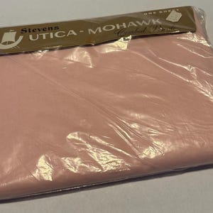 May include: A pink flat sheet in a clear plastic package. The packaging has a brown label with the text "Stevens UTICA - MOHAWK Combed Percale" and a logo of a sailboat. The sheet is folded and appears to be new.
