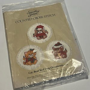 Könnte beinhalten: Ein Kreuzstich-Set für Weihnachtsschmuck. Das Set enthält drei runde Ornamente mit einer Katze, einem Bären und einem Hund in festlicher Kleidung. Der Text auf der Verpackung lautet "Something Special COUNTED CROSS STITCH Cat, Bear and Dog Ornaments". Jedes Ornament hat einen Durchmesser von 10 cm.