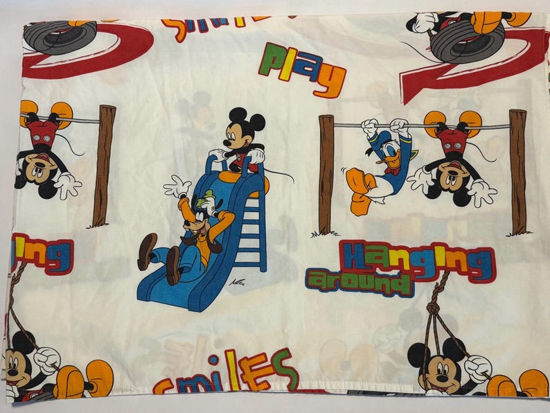 Puede incluir: S&aacute;bana blanca con un tema l&uacute;dico de Disney. Mickey Mouse, Goofy y el Pato Donald aparecen en ilustraciones coloridas. Tambi&eacute;n est&aacute;n presentes las palabras "Play", "Hanging Around" y "Smiles".