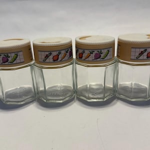 Peut inclure: Quatre pots à épices en verre transparent avec des couvercles beiges. Les couvercles ont une bande décorative avec des illustrations colorées de légumes. Les pots sont hexagonaux et vides.