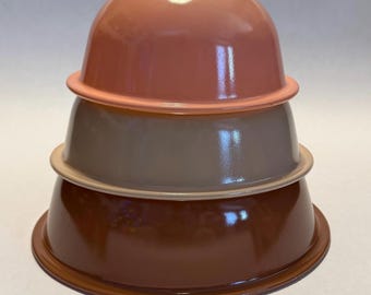 Puede incluir: Una pila de tres cuencos mezcladores vintage en tonos rosa, topo y marrón. Cada cuenco tiene una forma redondeada con un borde ligeramente ensanchado. Los cuencos están anidados, mostrando sus diferentes tamaños y colores. El acabado brillante refleja la luz.