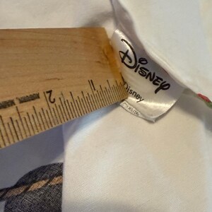 Puede incluir: Tela blanca con una etiqueta de Disney y una regla de madera. La regla mide la tela, mostrando una medida de aproximadamente 5 cm. La etiqueta de Disney es blanca con el logotipo de Disney y la palabra "Disney" impresa.