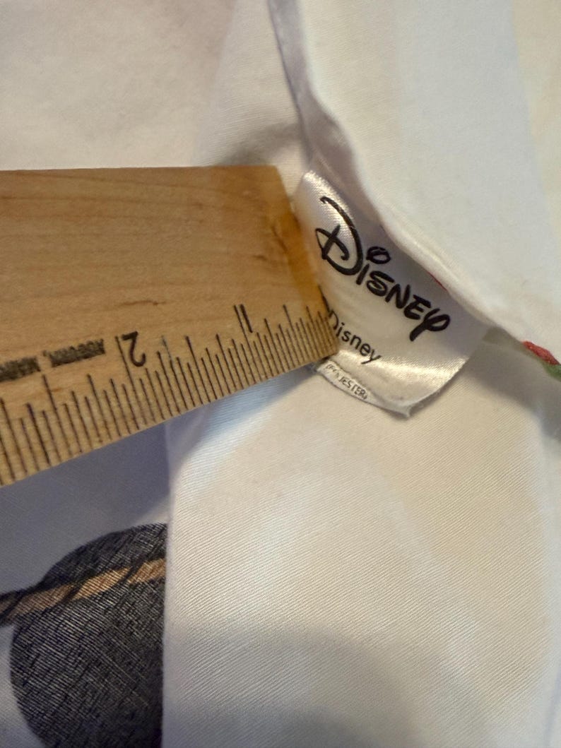 Puede incluir: Tela blanca con una etiqueta de Disney y una regla de madera. La regla mide la tela, mostrando una medida de aproximadamente 5 cm. La etiqueta de Disney es blanca con el logotipo de Disney y la palabra "Disney" impresa.
