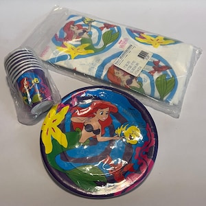 May include: A set of Little Mermaid themed party supplies, including a purple and blue paper plate featuring Ariel and Flounder, a stack of purple and white paper cups with Ariel, and a white tablecloth with Ariel and Flounder.