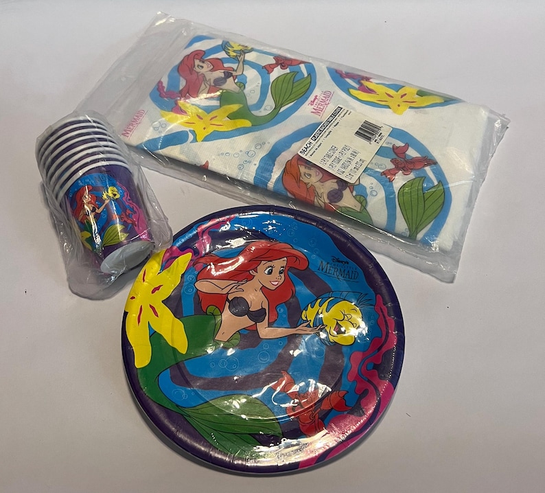 May include: A set of Little Mermaid themed party supplies, including a purple and blue paper plate featuring Ariel and Flounder, a stack of purple and white paper cups with Ariel, and a white tablecloth with Ariel and Flounder.
