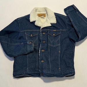 Puede incluir: Chaqueta vaquera azul oscuro con cuello y forro de borreguillo color crema. La chaqueta tiene dos bolsillos en el pecho, dos bolsillos laterales y cierre de botones. El logo de Wrangler es visible en un parche en el cuello.