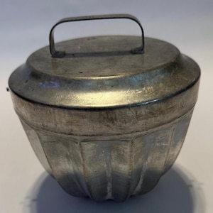 Könnte beinhalten: Eine silberfarbene Metall-Gugelhupfform mit Deckel und Griff. Die Form hat ein geriffeltes Design und einen abgerundeten Deckel mit einem kleinen Griff zum einfachen Anheben. Das Metall hat ein leicht gealtertes Aussehen.
