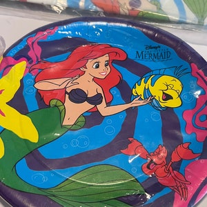 May include: A purple paper plate with a colourful design featuring Ariel from Disney's The Little Mermaid. The plate has a blue background with a swirling pattern and features Ariel, Flounder, and Sebastian. The text "Disney's The Little Mermaid" is printed on the plate.