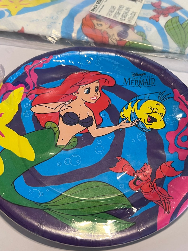May include: A purple paper plate with a colourful design featuring Ariel from Disney's The Little Mermaid. The plate has a blue background with a swirling pattern and features Ariel, Flounder, and Sebastian. The text "Disney's The Little Mermaid" is printed on the plate.