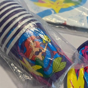 May include: A set of purple and white paper cups featuring a cartoon image of Ariel, the Little Mermaid, swimming with Flounder. The cups are packaged in a clear plastic bag.
