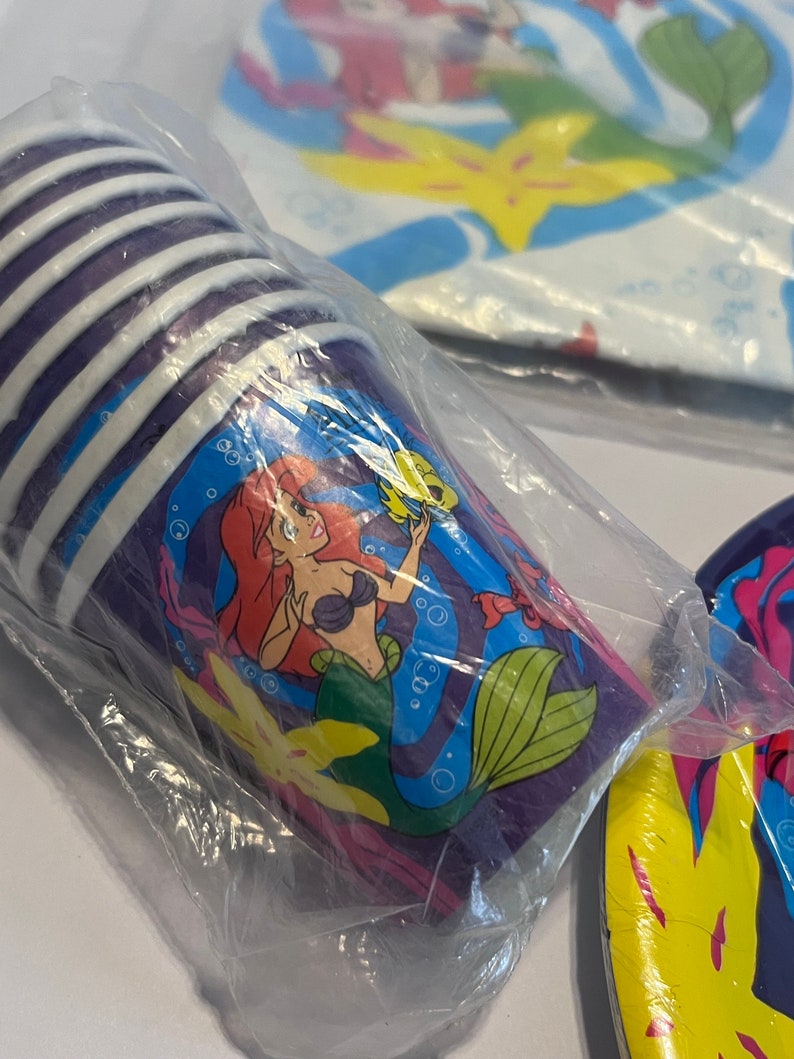 May include: A set of purple and white paper cups featuring a cartoon image of Ariel, the Little Mermaid, swimming with Flounder. The cups are packaged in a clear plastic bag.