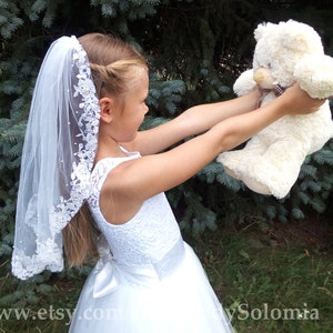 Puede incluir: Un velo de encaje blanco con intrincados bordados florales y pequeños detalles de perlas, combinado con un vestido blanco sin mangas con corpiño de encaje y una cintura de satén con lazo, sostenido junto a un oso de peluche color crema.