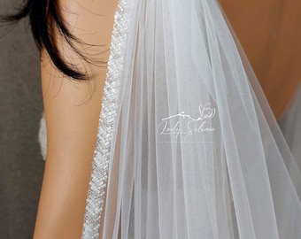 May include: A white tulle capelet featuring a silver sequin and bead embellished trim along the edge. The sheer fabric drapes from the shoulder, and a logo with the text Lady Salonia is visible on the center of the sheer white material.