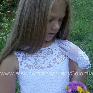 Puede incluir: Un vestido blanco sin mangas con un corpi&ntilde;o de encaje con patr&oacute;n floral y una cintura de sat&eacute;n lisa. La prenda se combina con guantes blancos transparentes hasta la mu&ntilde;eca que presentan un intrincado bordado de encaje blanco en los pu&ntilde;os.