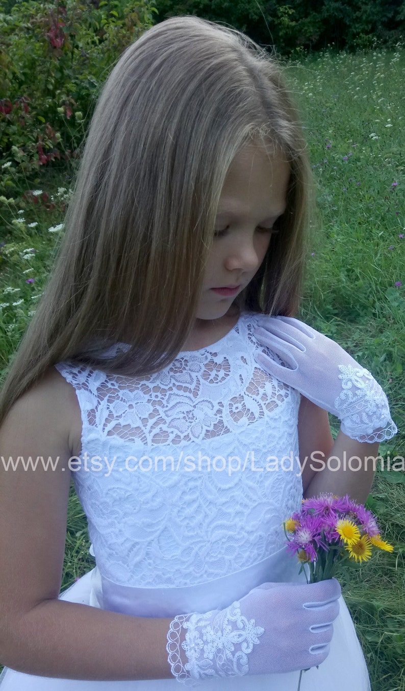 Puede incluir: Un vestido blanco sin mangas con un corpi&ntilde;o de encaje con patr&oacute;n floral y una cintura de sat&eacute;n lisa. La prenda se combina con guantes blancos transparentes hasta la mu&ntilde;eca que presentan un intrincado bordado de encaje blanco en los pu&ntilde;os.