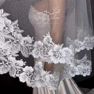May include: A white wedding veil featuring a shimmering, glitter-infused tulle fabric and an intricate floral lace border. The lace displays detailed white embroidered flowers and leaves along the scalloped edge. The text Lady Solomia is visible.