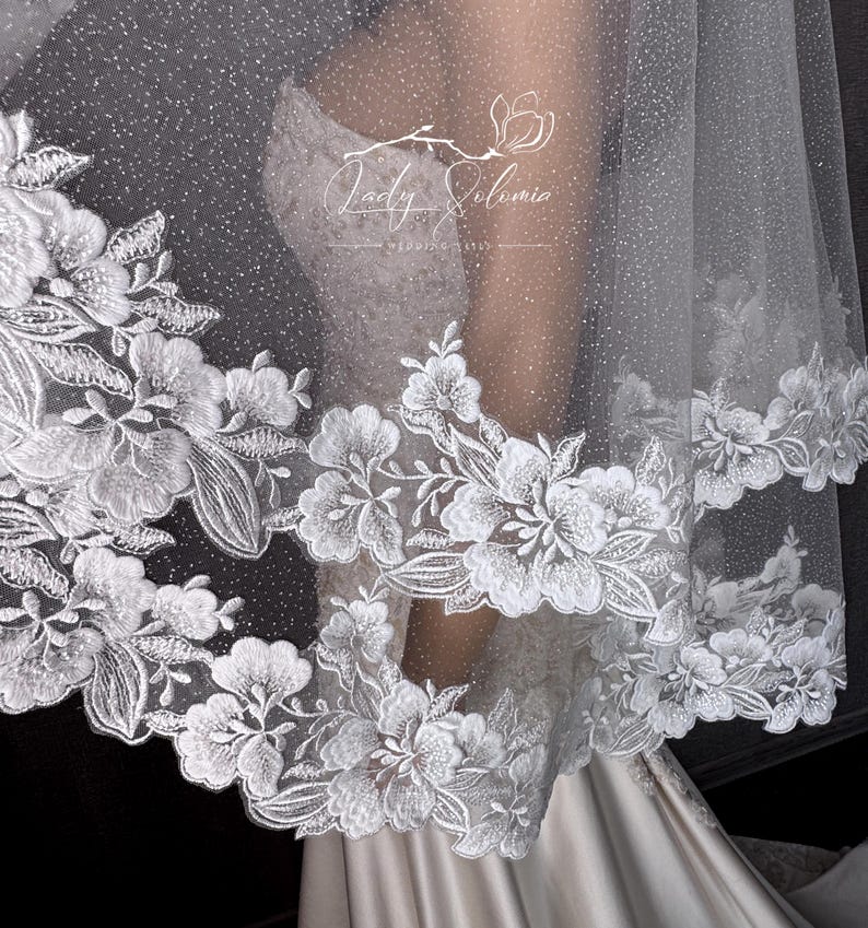 May include: A white wedding veil featuring a shimmering, glitter-infused tulle fabric and an intricate floral lace border. The lace displays detailed white embroidered flowers and leaves along the scalloped edge. The text Lady Solomia is visible.