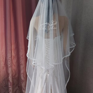 Puede incluir: Un velo de novia blanco de dos niveles con un acabado de purpurina sutil y un borde festoneado delicado. El velo est&aacute; drapeado sobre un vestido sin tirantes con espalda de cors&eacute; con cordones. El texto Lady Solomia est&aacute; impreso en blanco.
