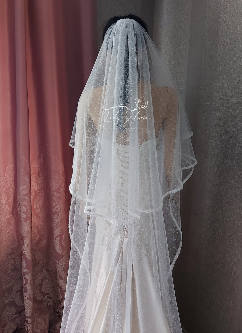 Puede incluir: Un velo de novia blanco de dos niveles con un acabado de purpurina sutil y un borde festoneado delicado. El velo est&aacute; drapeado sobre un vestido sin tirantes con espalda de cors&eacute; con cordones. El texto Lady Solomia est&aacute; impreso en blanco.