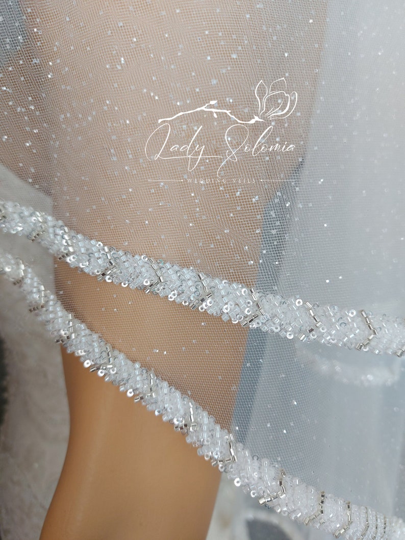 Puede incluir: Un velo de novia blanco transparente con un acabado de purpurina fina en la tela de tul. Los bordes est&aacute;n adornados con un borde decorativo de cuentas de semillas blancas y cuentas de tubo met&aacute;licas plateadas. El texto Lady Solomia Wedding Veils es visible.