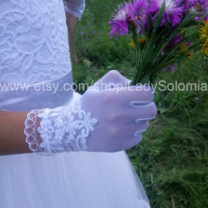 Puede incluir: Guantes de malla blanca con intrincados bordados de encaje floral en la mu&ntilde;eca, combinados con un vestido de encaje blanco. Los guantes se sostienen junto a un ramo de flores silvestres moradas y amarillas. El texto www.etsy.com/shop/LadySolomia es visible.