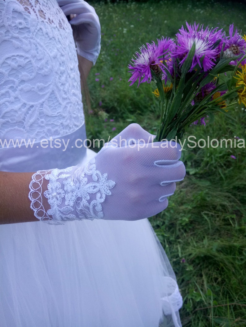 Puede incluir: Guantes de malla blanca con intrincados bordados de encaje floral en la mu&ntilde;eca, combinados con un vestido de encaje blanco. Los guantes se sostienen junto a un ramo de flores silvestres moradas y amarillas. El texto www.etsy.com/shop/LadySolomia es visible.