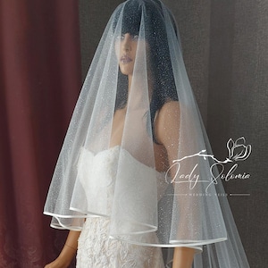 May include: A white tulle wedding veil with a single layer and a satin edge. The veil is decorated with small, sparkly dots. The veil is shown on a mannequin.
