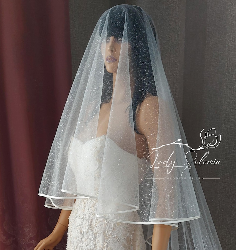 May include: A white tulle wedding veil with a single layer and a satin edge. The veil is decorated with small, sparkly dots. The veil is shown on a mannequin.