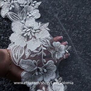 May include: A white lace trim featuring intricate floral embroidery with silver thread accents and clear sequins on a sheer mesh base. The fabric displays a repeating pattern of blossoms and leaves. Text reads www.etsy.com/shop/LadySolomia.