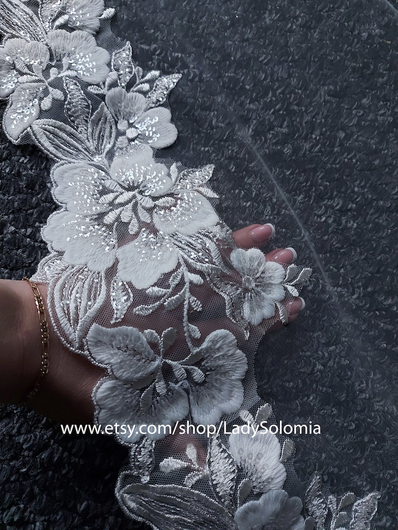 May include: A white lace trim featuring intricate floral embroidery with silver thread accents and clear sequins on a sheer mesh base. The fabric displays a repeating pattern of blossoms and leaves. Text reads www.etsy.com/shop/LadySolomia.