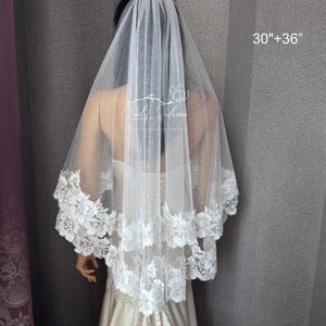 May include: A two-tier white bridal veil featuring a shimmering glitter tulle fabric and a scalloped floral lace trim along the edges. The veil is labeled with the text 30"+36" indicating its length. The design includes intricate white floral embroidery.
