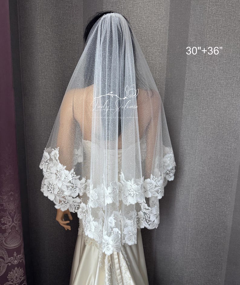 May include: A two-tier white bridal veil featuring a shimmering glitter tulle fabric and a scalloped floral lace trim along the edges. The veil is labeled with the text 30"+36" indicating its length. The design includes intricate white floral embroidery.