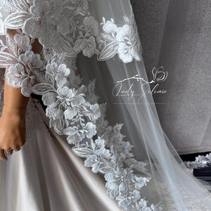 May include: A white wedding veil featuring intricate floral lace embroidery along the edges. The sheer tulle fabric drapes over a smooth, light-colored gown. A logo with the text Lady Solomia Wedding Veils is visible on the sheer fabric.