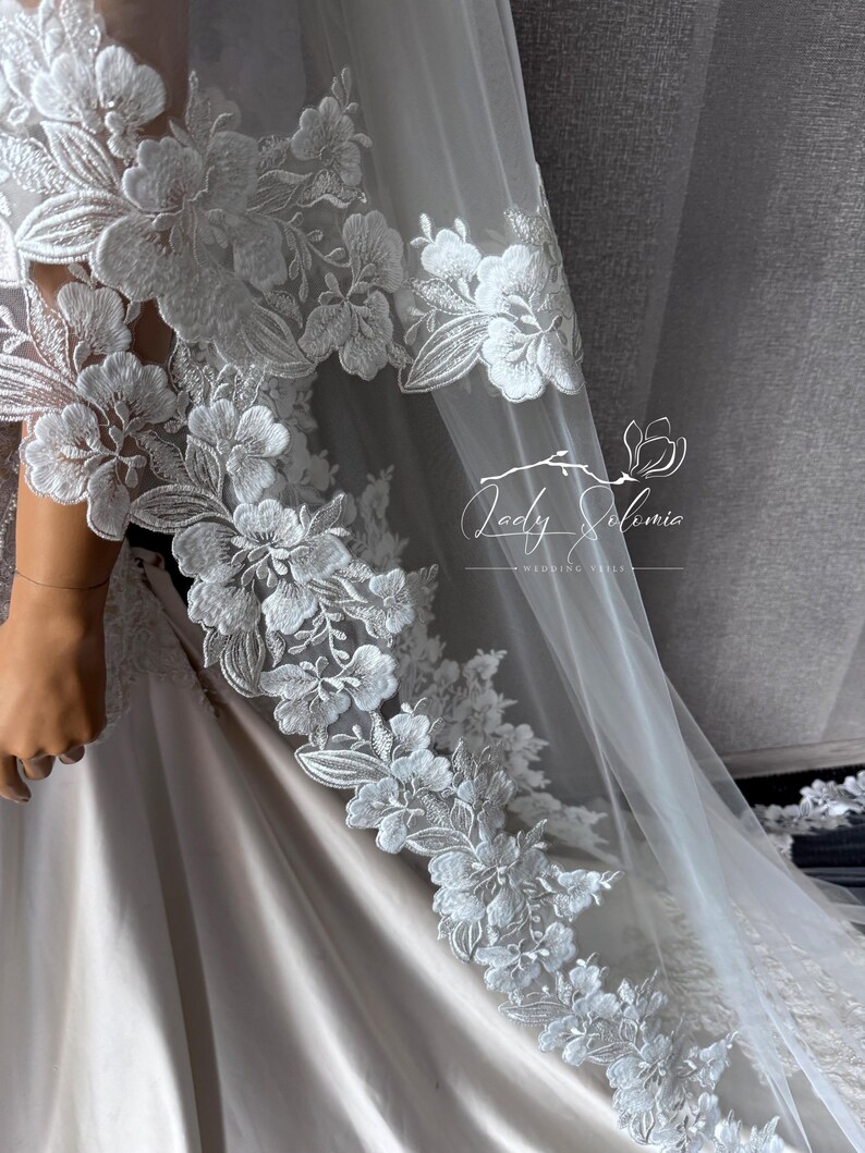 May include: A white wedding veil featuring intricate floral lace embroidery along the edges. The sheer tulle fabric drapes over a smooth, light-colored gown. A logo with the text Lady Solomia Wedding Veils is visible on the sheer fabric.