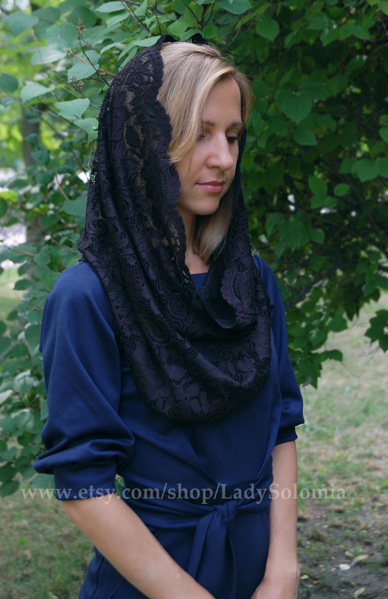May include: A black lace head covering with a floral pattern. The lace is draped over a woman's head and shoulders. She is wearing a dark blue dress.