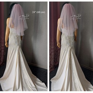 May include: A pale pink, two-tier tulle wedding veil measuring 24 inches in length. The veil is draped over a mannequin wearing a cream-colored, lace-detailed gown with a corset-style lace-up back closure and a long, flowing train.