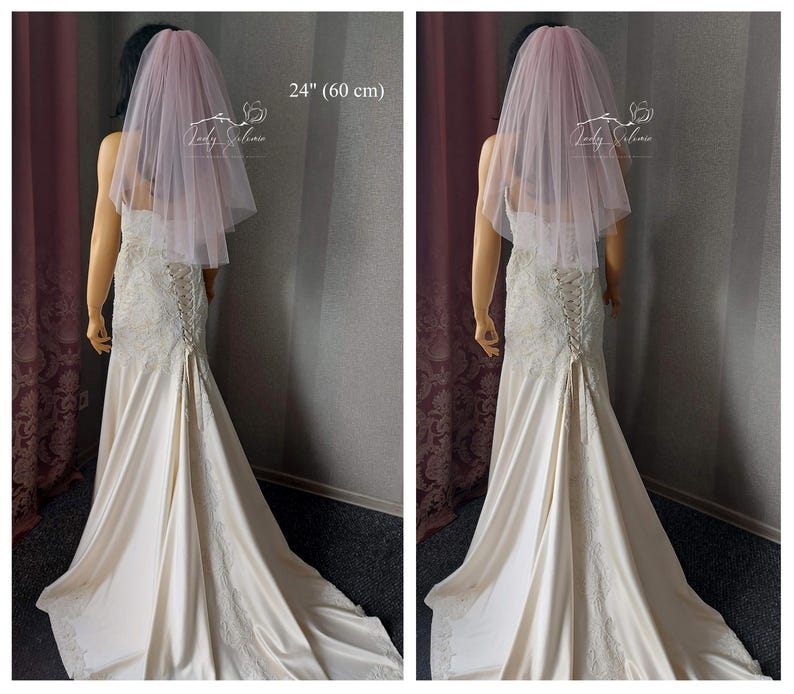 May include: A pale pink, two-tier tulle wedding veil measuring 24 inches in length. The veil is draped over a mannequin wearing a cream-colored, lace-detailed gown with a corset-style lace-up back closure and a long, flowing train.