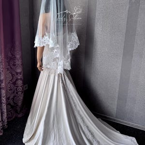 May include: A floor-length ivory wedding dress with a satin finish and a long train featuring intricate white floral lace embroidery along the hem and back. A sheer white tulle veil with matching floral lace trim rests over the shoulders.