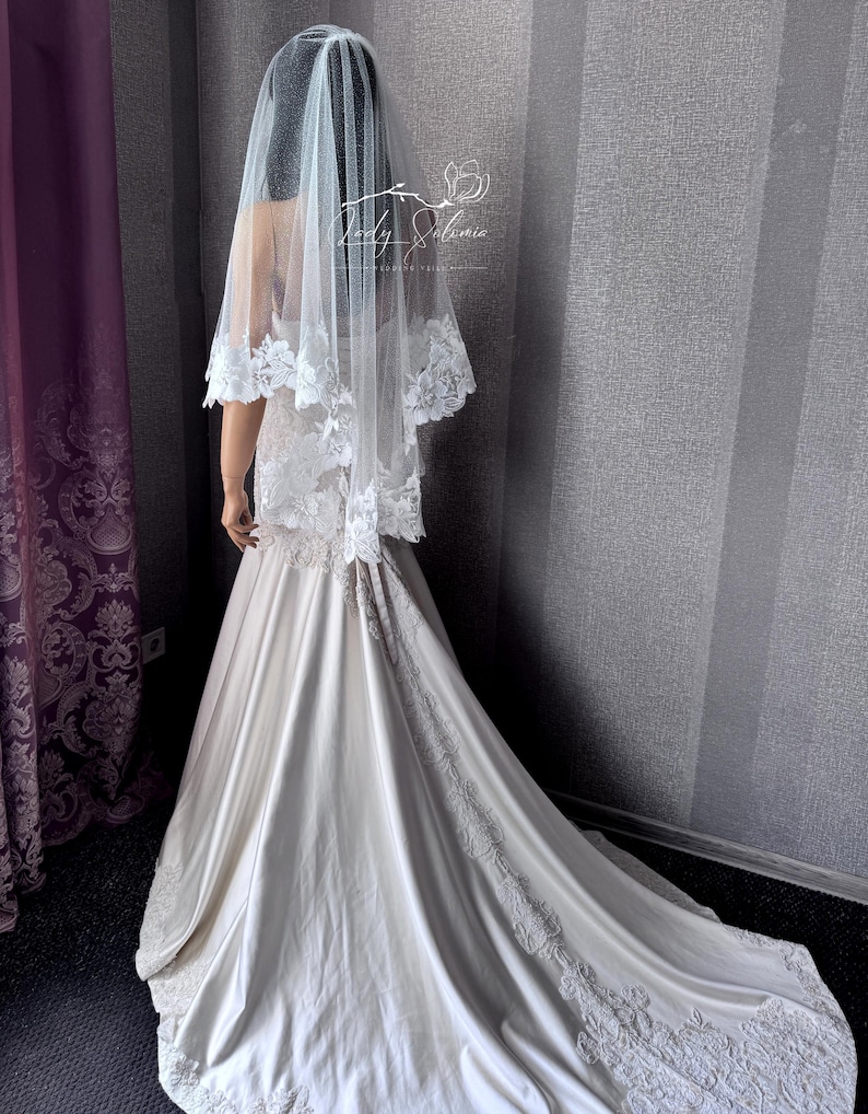 May include: A floor-length ivory wedding dress with a satin finish and a long train featuring intricate white floral lace embroidery along the hem and back. A sheer white tulle veil with matching floral lace trim rests over the shoulders.