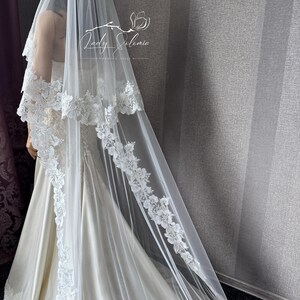 May include: A long white wedding veil featuring intricate floral lace embroidery along the edges, draped over a mannequin wearing a satin gown. The veil includes the text Lady Solomia Wedding Veil printed in white near the top center section.