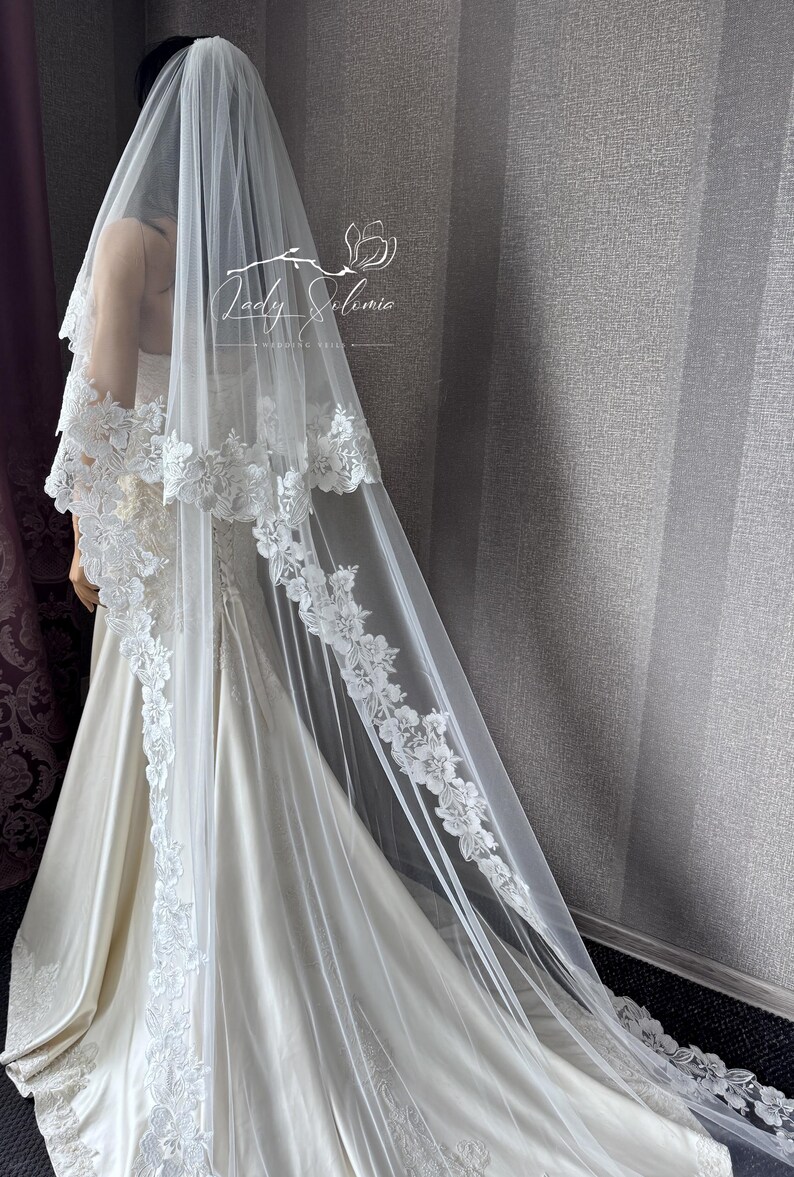 May include: A long white wedding veil featuring intricate floral lace embroidery along the edges, draped over a mannequin wearing a satin gown. The veil includes the text Lady Solomia Wedding Veil printed in white near the top center section.