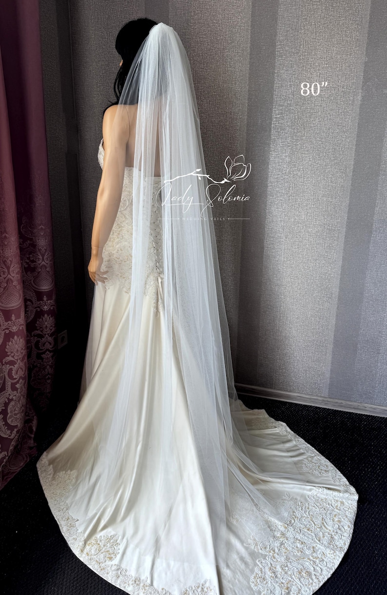 May include: A long, sheer white wedding veil measuring 80 inches in length, draped over a floor-length ivory gown with lace detailing at the hem. The veil is made of fine, translucent tulle fabric and features a simple, unadorned edge design.